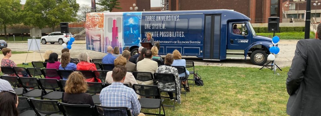 Crowd sitting at presentation outside in front of UI Ride Shuttle Bus
